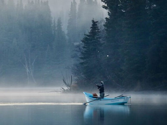 A person standing in a boat along the Kenai River casts a fly fishing line into the misty water.