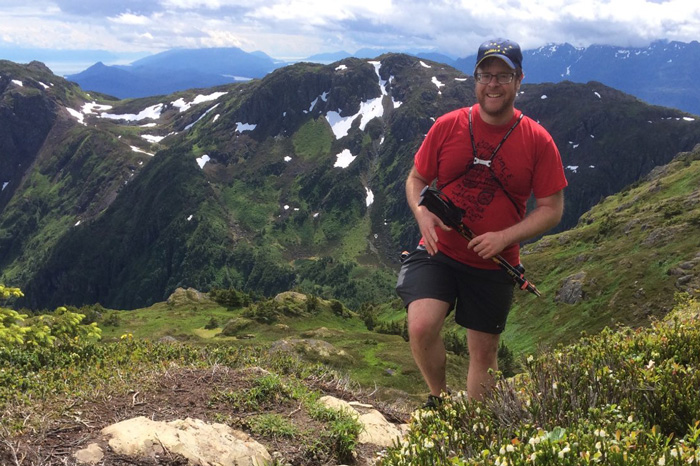 Andy Miller hikes up a mountain with a stunning mountain vista in the background and wild flowers in the foreground.