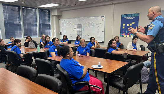 Jr. DA program participants sit in a classroom listening to a police officer who stands at the front of the classroom.