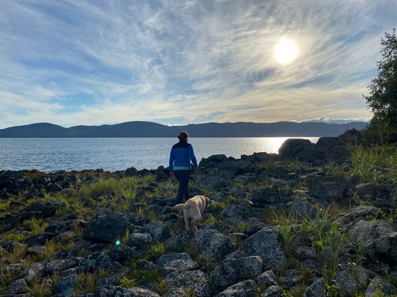 A hiker and her dog walks across a rocky beach. Wispy clouds are in the sky.