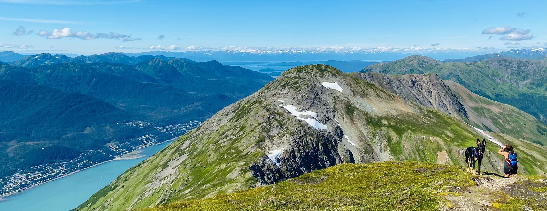 A hiker and her dog stand on Mt. Roberts with a view of the mountains and water below.