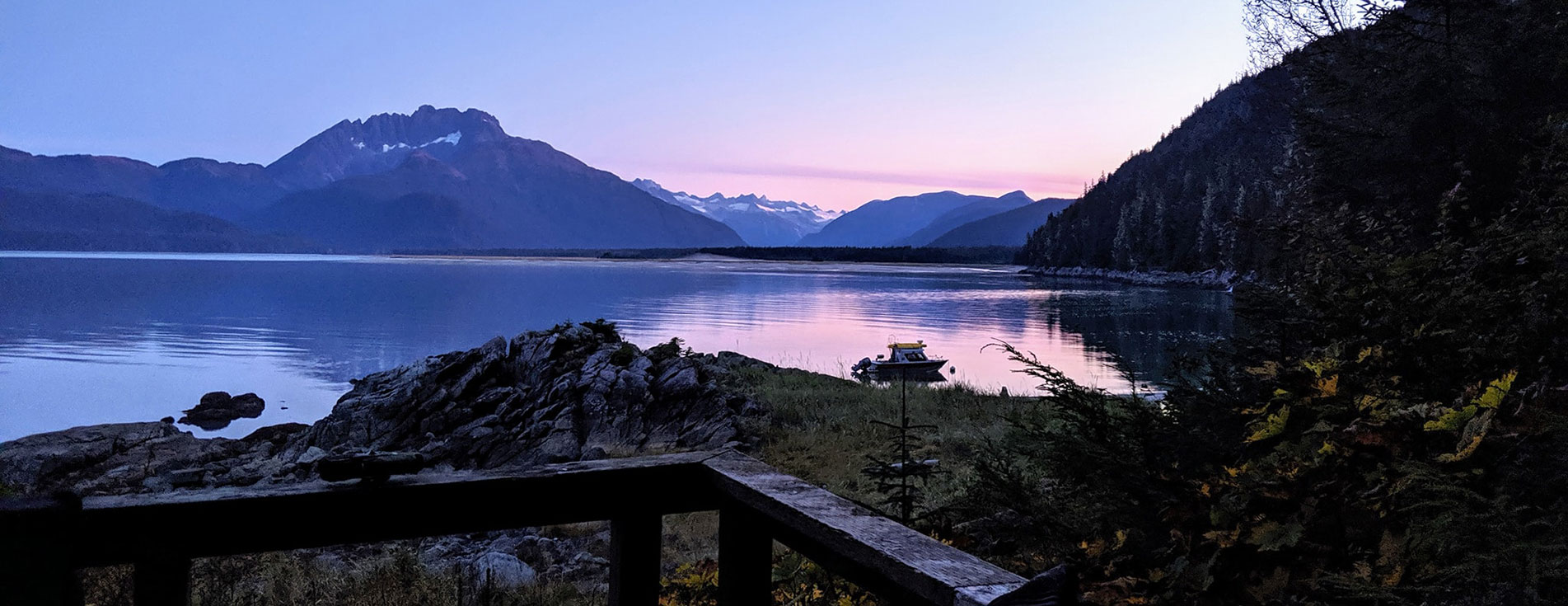 The sun sets behind the mountians across the water as seen from a beach-side railing. A small boat is anchored in the water.