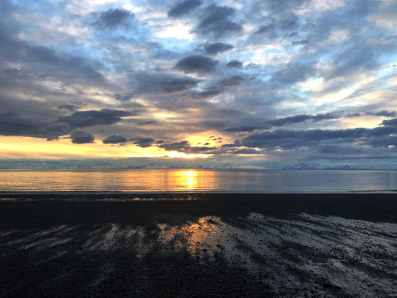 The sun sets behind the mountains across the water along the beaches of the Kenai River.