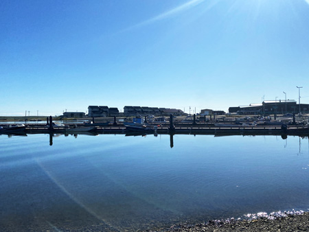 Small boats line the harbor in Kotzebue. Blue skies reflect on calm water. Buildings can be seen in the bakground