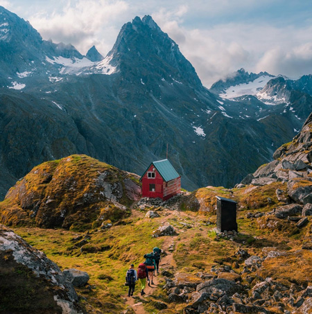 Four backpackers approach a small red cabin, Mint Hut with mountain peaks in the background